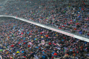 Eine große Menschenmenge sitzt und steht in einem Stadion mit Geländern und Texttafeln im Hintergrund.