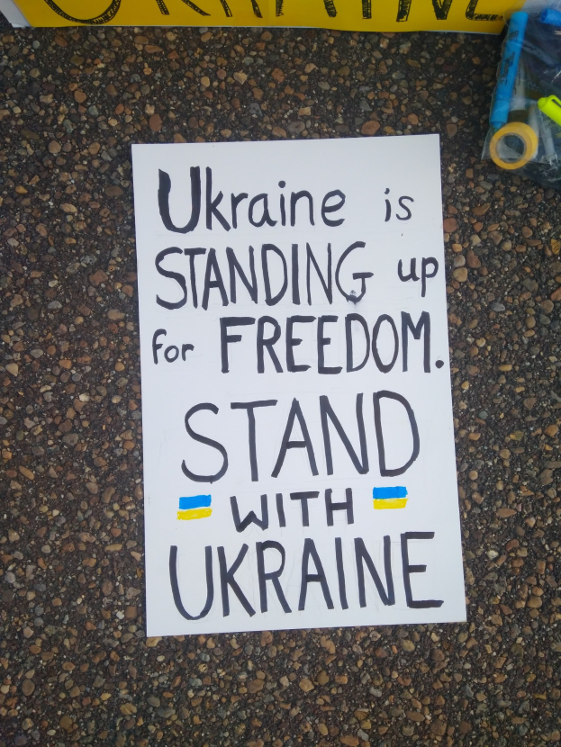 Weißer Zettel mit "Ukraine steht für Freiheit, steht mit Ukraine" Text auf dem Boden vor einem gelben Banner, mit einer Plastikabdeckung mit verschiedenen Gegenständen rechts.