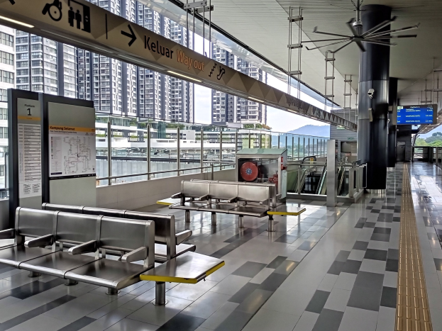 Leerer Flughafen Wartebereich mit mehreren Bänken, Informationsanzeigen, Säulen, Deckenventilator und Leuchten; Hintergrund zeigt Gebäude, Bäume, Wasser und einen klaren blauen Himmel.