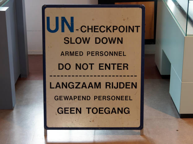 Ein Schild auf dem Boden lautet "UN-Checkpoint Slow Down Armed Personnel - Do Not Enter", mit einem Glastisch mit verschiedenen Gegenständen darauf rechts und einer Wand im Hintergrund.
