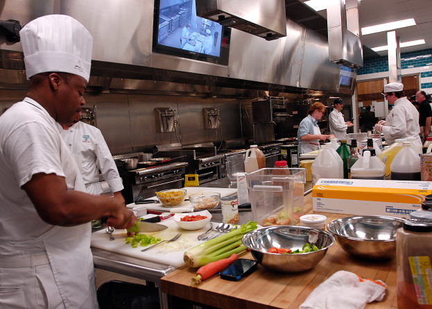 Eine Gruppe von Köchen in einer Küche, die auf einem Schneidbrett Lebensmittel zubereiten, wobei ein Koch in der foreground in einem weißen Kleid und einer Mütze steht und ein Messer hält und von Utensilien, Schüsseln, Gemüse, einem Mobiltelefon, Flaschen und anderen Gegenständen umgeben ist, mit einem Fernseher und Deckenleuchten im Hintergrund.