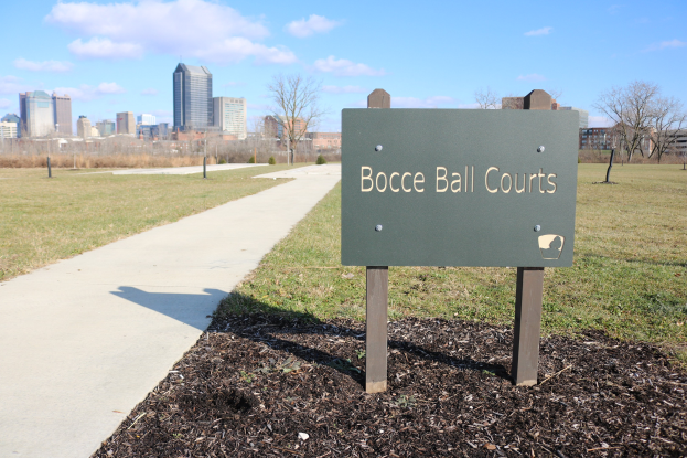 Ein Schild am Boden zeigt Boccia-Plätze in Indianapolis, Indiana, mit Gras, einem Weg, Bäumen, Pfählen, einem Zaun, Gebäuden und einem bewölkten Himmel im Hintergrund.