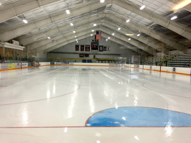 Große Indoor-Eisbahn mit Deckenbeleuchtung, Werbetafeln, einer Rückwand, Treppen mit Geländern und einigen Menschen, die einer Hockey-Anlage ähneln.