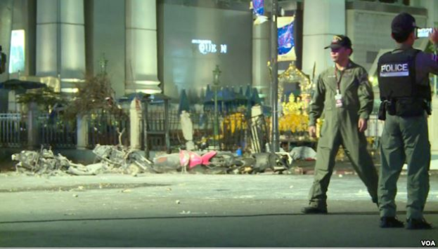 Zwei Polizisten in Uniform gehen neben Schutt eine Straße entlang, mit Gebäuden, Säulen und anderen Objekten im Hintergrund und Text am unteren Bildrand.