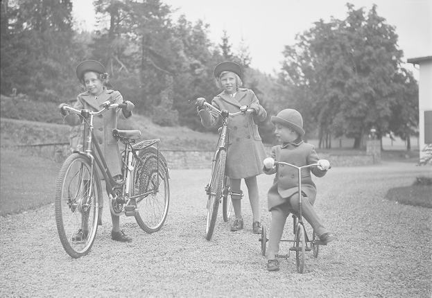 Drei Kinder in Mänteln und Hüten fahren Fahrräder in einem Park mit Bäumen und einem Haus im Hintergrund, aufgenommen in Schwarzweiß.