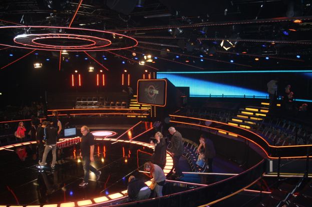 Gruppe von Menschen auf einer hell erleuchteten Bühne mit Stühlen und einem Bildschirm im Hintergrund, wahrscheinlich von einer Fernsehsendung.