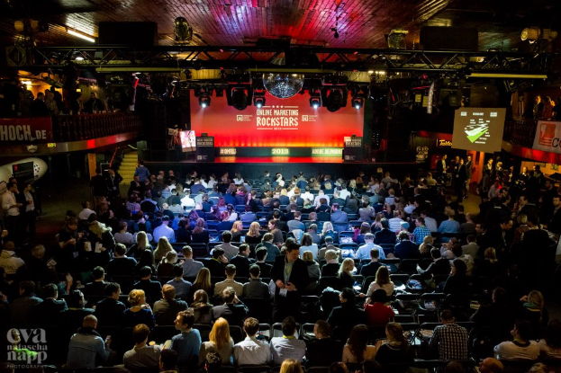 Eine große Menschenmenge sitzt auf Stühlen vor einer Bühne mit Bildschirmen und Plakaten, beleuchtet von Scheinwerfern und umgeben von Absperrungen, voller Erwartung auf den Beginn eines Online-Marketing-Rockstars-Events, mit einem Wasserzeichen in der linken unteren Ecke.