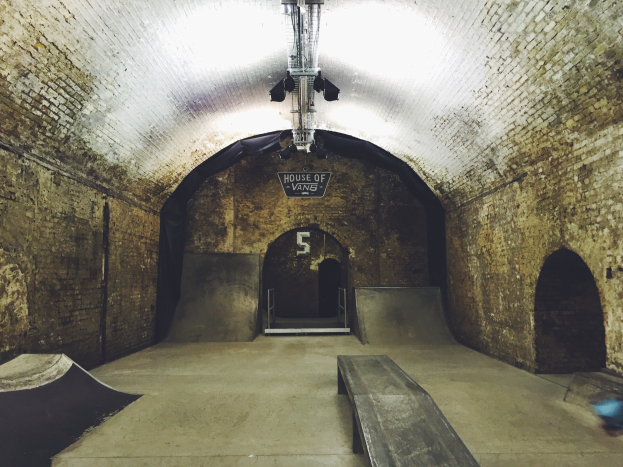 Ein Skatepark mit Backsteinmauer, Rampe, Bank, Schild und Deckenbeleuchtung.