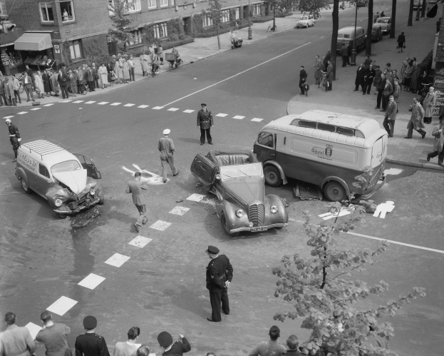 Schwarz-weiß-Szene eines Autounfalls an einer Straßenkreuzung mit mehreren Fahrzeugen, darunter ein Van, und einer Gruppe von Menschen in der Nähe, mit Gebäuden, Bäumen, Pfählen und Schildern im Hintergrund.