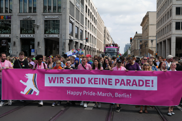 Eine Gruppe von Menschen marschiert auf einer Straße in Berlin mit einem pinken Banner, auf dem "Happy Pride March" steht, mit Gebäuden, Laternenmasten und Verkehrszeichen an der Straße unter einem bewölkten Himmel.