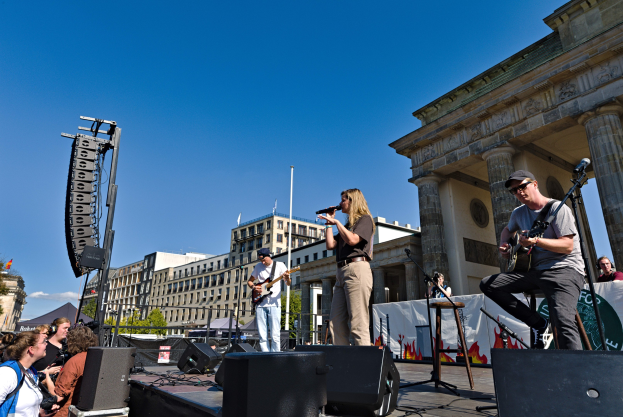 Eine Gruppe von Menschen, die auf einer Bühne vor dem Brandenburger Tor in Berlin Musik spielen, mit Lautsprechern und Equipment drumherum, unter einem klaren blauen Himmel.