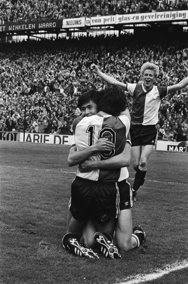 Drei Fußballspieler umarmen sich auf dem Boden nach einem Tor, mit jubelnder Menge und schwenkenden Fahnen in einem schwarz-weißen Stadion Setting.