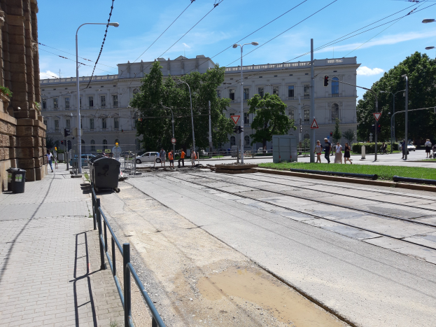Eine Stadtstraße mit einer durch sie verlaufenden Bahnschiene, umgeben von Gebäuden, Straßenlaternen, Strommasten, Stromkabeln, Kraftfahrzeugen, Fußgängern, Mülltonnen, Absperrpollern, Büschen, Bäumen und einem bewölkten Himmel.