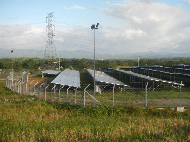 Ein Solarpanelfeld mit einem Zaun im Vordergrund, umgeben von Gras und Pflanzen, Bäumen, Übertragungsmasten mit Drähten und einem bewölkten Himmel im Hintergrund.