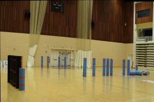 Großer Turnsaal mit einem Basketballfeld, Geräteracks auf der rechten Seite, Vorhänge und ein Bildschirm an der Wand im Hintergrund.