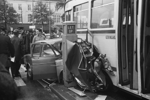 Schwarzes und weißes Bild eines Busses, der auf der Seite der Straße in ein Auto gekracht ist, mit Menschen, die sich darum versammelt haben, und Bäumen und Gebäuden im Hintergrund.