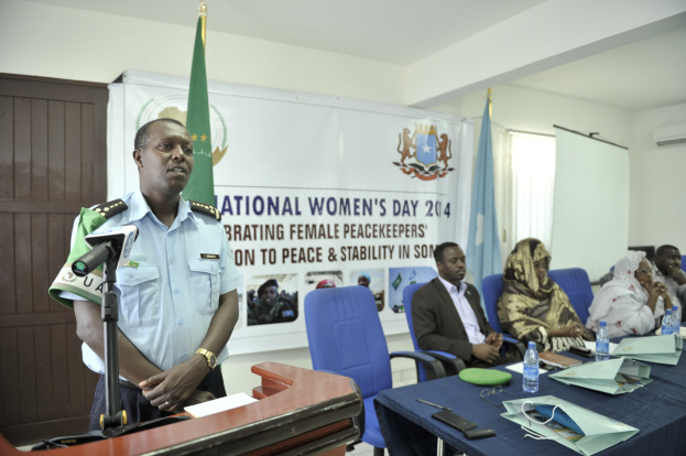 Ein als Chef de Cabinet des Nationalen Frauentags 2014 identifizierter Mann steht an einem Rednerpult mit einem Mikrofon und einem Blatt Papier und spricht vor einem Publikum, das auf Stühlen sitzt, mit verschiedenen Gegenst"anden auf dem Tisch und Fahnen, einem Banner, einer T"ur, einer Klimaanlage, einem Fenster und einer Wand im Hintergrund.