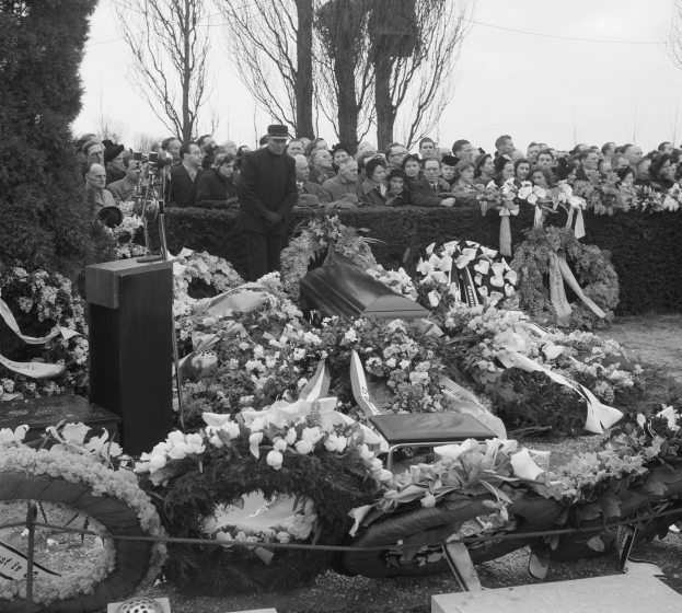 Schwarzer und weißer Trauerzug mit einem Mann in einer Mütze, der auf einem Podium spricht und von Blumen, Stühlen und einer Menge umgeben ist, mit Bäumen und einem klaren Himmel im Hintergrund.