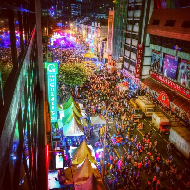 Eine pulsierende Karnevalsszene mit einer großen Menschenmenge auf der Straße, umgeben von Zelten, Geschäften, Gebäuden und Bannern, mit hell erleuchteten Bereichen im Hintergrund.