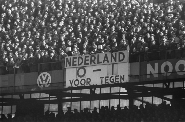 Ein Schwarz-Weiß-Foto einer großen Menschenmenge in einem Stadion, mit einer zentralen Tafel, die Text anzeigt, und ein paar Individuen, die unten stehen.