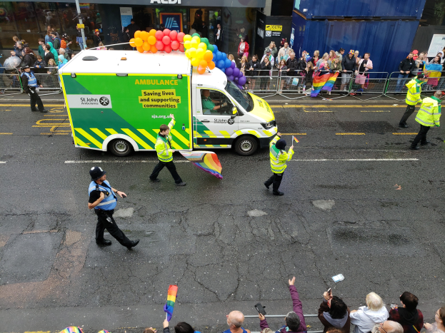Eine Gruppe von Menschen in grünen Jacken, einige mit Fahnen, geht eine Straße entlang mit einem Ballon-dekorierten Krankenwagen im Hintergrund, Gebäude mit Glastüren und Namensschildern weiter hinten sichtbar.