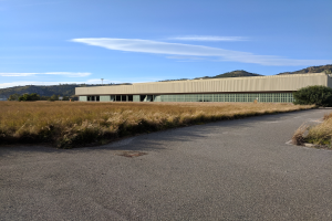 Leere Straße, die zum Hauptquartier des San Francisco International Airport führt, ein großes Gebäude auf einem Feld umgeben von Pflanzen und Bäumen, mit Hügeln und einem bewölkten Himmel im Hintergrund.