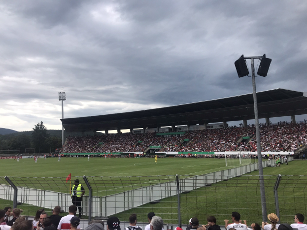 Große Zuschauermenge bei einem Fußballspiel in einem bewölkten, offenen Stadion mit beleuchtetem Feld, umgeben von einem Zaun, Bäumen und Hügeln.