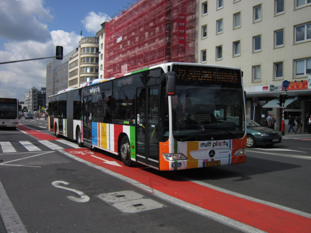 Eine Stadtbus fährt eine Straße mit hohen Gebäuden entlang, mit Menschen auf dem Gehweg, Ampeln und Texttafeln, unter einem bewölkten Himmel.