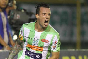 Ein Fussballer in grün-weißer Uniform mit Tattoos auf dem Arm feiert nach einem Tor, mit unscharfen Figuren und einer Tafel im Hintergrund.