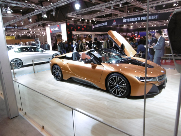 Ein stylischer schwarzer BMW i8 Roadster mit Chromakzenten, langer Motorhaube und niedriger Karosserie, präsentiert auf der IAA in Frankfurt umgeben von formell gekleideten Gästen.