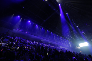 Große Menschenmenge in einem abgedunkelten Konzertsaal gegenüber einer Bühne mit Deckenleuchten und einem Bildschirm auf der rechten Seite.