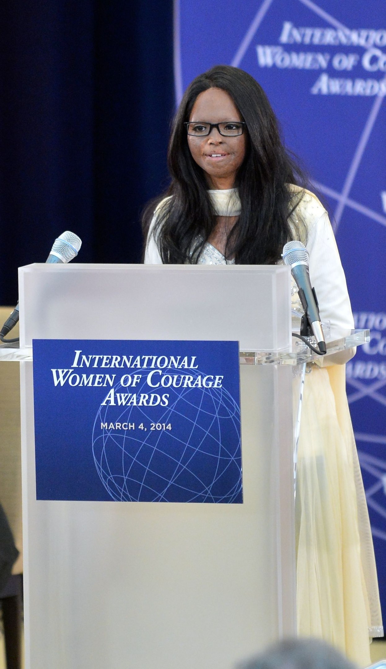 Frau in weißem Kleid und Brille spricht an einem Pult mit zwei Mikrofonen und einer Tafel, während der International Women of Courage Awards 2014, mit einem blauen Banner im Hintergrund.