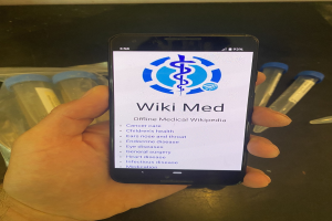 Eine Person hält ein Smartphone in der Hand, auf dem die WikiMed-App geöffnet ist, mit Reagenzgläsern und anderen Gegenständen auf dem Tisch davor.