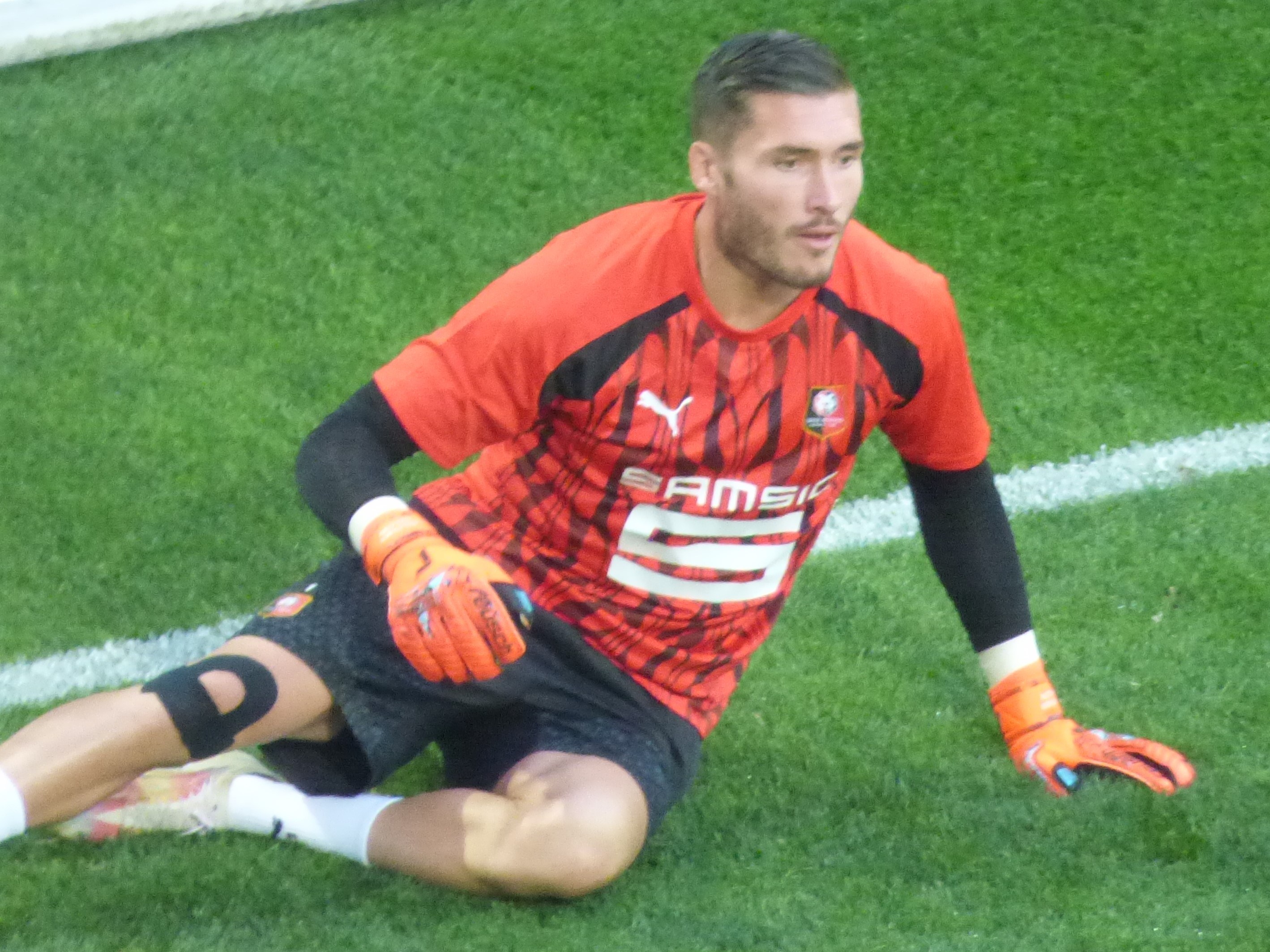 Ein Fußballer in rotem T-Shirt, schwarzen Shorts, weißen Socken und orangen Baseball-Handschuhen sitzt auf dem Boden mit den Händen auf den Knien.