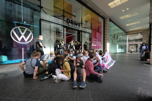 Menschen sitzen auf dem Boden vor einer Volkswagen-Niederlassung und halten ein Transparent.