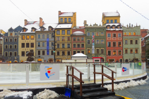 Eislaufbahn in einem Stadtplatz umgeben von Gebäuden, Treppen, Geländern, Informationsschildern, schneebedeckter Fläche, Laternen, Bäumen und einem Himmel als Hintergrund.