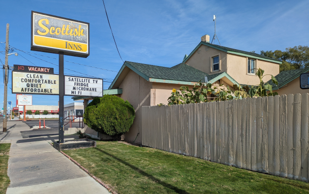 Ein schottisches Motel in San Francisco, Kalifornien, mit einem Haus mit Dach und Fenstern, einem hölzernen Zaun, Pflanzen, Gras, Bäumen, Schildern, Strommasten mit Drähten, einer Straßenlaterne, Fahrzeugen auf der Straße und einem bewölkten Himmel.