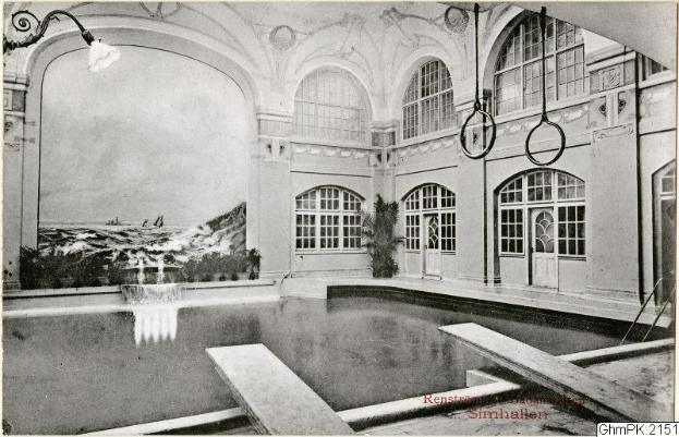 Ein altes Schwarz-Weiß-Foto eines großen Hallenbades in einem Gebäude, umgeben von einer Wand mit Glasfenstern und -türen, mit Pflanzen und einer Deckenlampe. Unten steht "Rensburg, Deutschland - Schwimmbad".