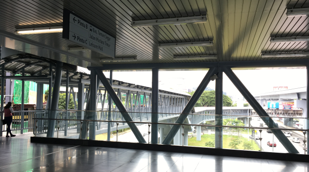 Innenansicht eines Flughafen-Terminals mit Menschen, Geländern, Säulen, Informationsanzeigen, Deckenleuchten und einer Außenansicht einer Straße, Brücke, Vegetation, Gebäuden und Himmel.