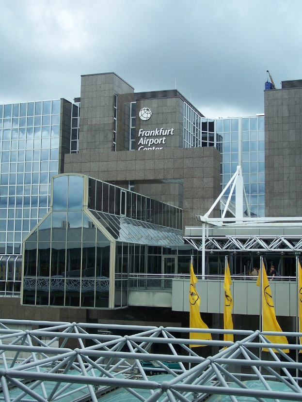 Frankfurt Airport building with glass walls, text, yellow flags, and iron rods under a cloudy sky.