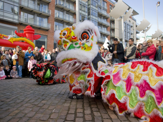 Ein lebendiges chinesisches Neujahrsfest in Amsterdam mit einem Löwendance im Vordergrund, umgeben von einer Menschenmenge, mit Gebäuden, Laternenmästen und einem klaren blauen Himmel im Hintergrund.