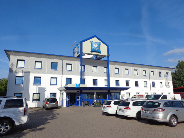 Ein Gebäude mit einem Schild, das "ibis budget frankfurt airport" liest, geparkte Autos, Topfpflanzen, eine Straßenlaterne, Bäume und einen bewölkten Himmel.