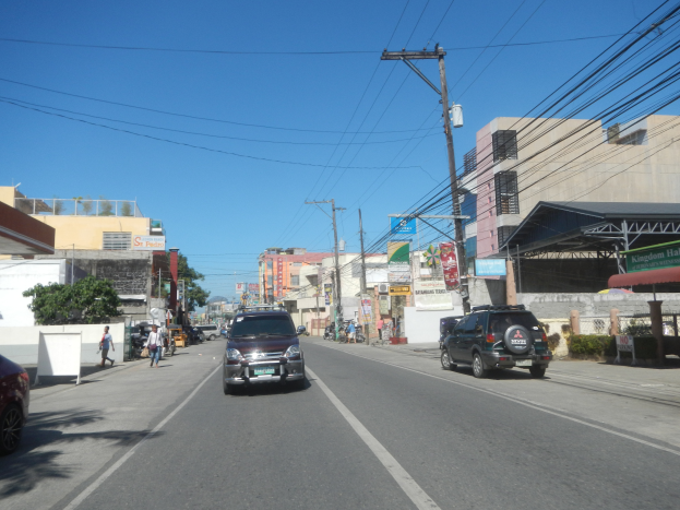 Eine Stadtstraße mit fahrenden Autos, Fußgängern auf den Gehwegen, Strommasten, Gebäuden mit Fenstern, Bäumen und einem klaren blauen Himmel.