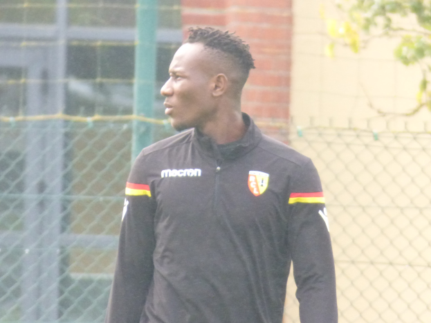 Ein Mann In Einer Schwarzen Jacke Steht Auf Einem Fussballfeld, Umgeben Von Einem Zaun Und Eines Baumes, Und Sieht konzentriert Aus.