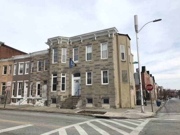 Reihe von Häusern an einer Straßenecke mit Fahrzeugen, Bäumen und einem bewölkten Himmel im Hintergrund.