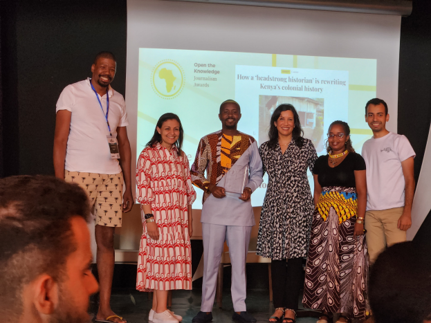 Eine Gruppe von Menschen auf einer Bühne, die lächeln und für ein Foto posieren, mit einem Publikum davor und einem Bildschirm im Hintergrund, der 'Open the Knowledge Journalism Awards 2019' anzeigt.