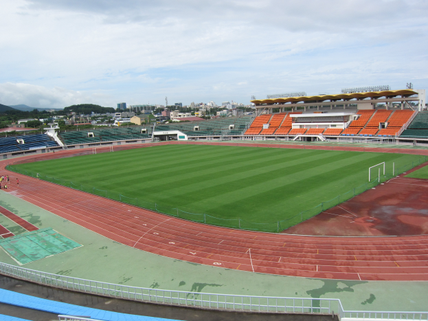 Ein großes Stadion mit einem Fußballfeld in der Mitte, umgeben von Gebäuden, Bäumen und Hügeln unter einem klaren blauen Himmel, mit wenigen Menschen und saftig grünem Gras auf dem Boden.