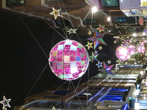 Eine nächtliche Straßenansicht mit zahlreichen Lichtern und Dekorationen, die einen großen Diskokugel an einem Gebäude hängen, schaffen eine festliche Atmosphäre.