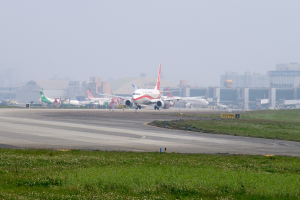 Flughafenvorfeld mit mehreren geparkten Flugzeugen, grünem Umfeld, Gebäuden im Hintergrund und einem klaren blauen Himmel.
