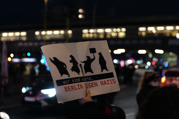 Eine Person hält ein Schild mit der Aufschrift "Berlin gegen Nazis" vor einer Menge und Fahrzeugen auf einer Straße, mit einem beleuchteten Gebäude im Hintergrund.